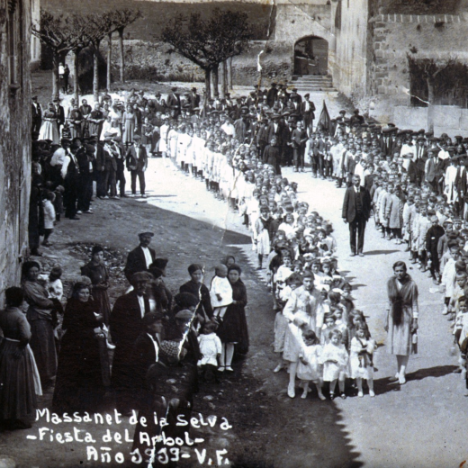 Festa de l'arbre de l'any 1919. Els col·legis desfilant per la plaça amb els mestres Srta. Angela Baró i Pere Cantenys. Al fons es pot apreciar el típic porxet de la rectoria i la pared que tancava la plaça, avui tot desaparegut per donar pas al desenvolupament urbanístic