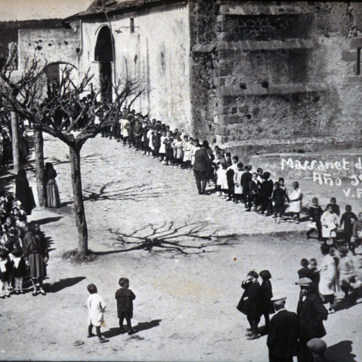 La mainada de les escoles amb els respectius mestes, Pere Cantenys i Angela Baró, sortint de l'Església després de celebrar-se un acte religiós i encaminant-se cap el nou grup escolar per participar en l'acte de la seva inaguració oficial. Destaca en aquesta foto el porxet de la rectoria i el molí de vent que durant molts anys serví per pujar l'aigua a la rectoria i regar l'hort i el seu jardí.
