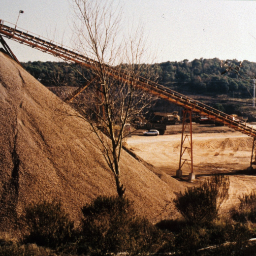 La pedrera de Can Figueras, un piló de pedra capulada i la cinta transportadora; al fons el turó d'on es treu la Roca.
