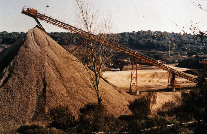 La pedrera de Can Figueras, un piló de pedra capulada i la cinta transportadora; al fons el turó d'on es treu la Roca.