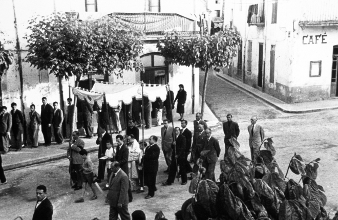 La processó de Corpus al seu pas per la plaça de l'Església. El rector és Mnh Conrad Ruhí i els portadors del Tàlem són en Joan Munsó, Josep Reyner, Martí Rossell, Josep Bancells i Eliodoro Palomo. Les autoritats són Narcís Planas, alcalde, Joan Viader, jefe de la Falange, Martí Vilella, jutge, Francesc Bosch, regidor, Joan Artigas, regidor, Josep Aregay, sindicat i el Sr. Joan Negre, metge titular.
