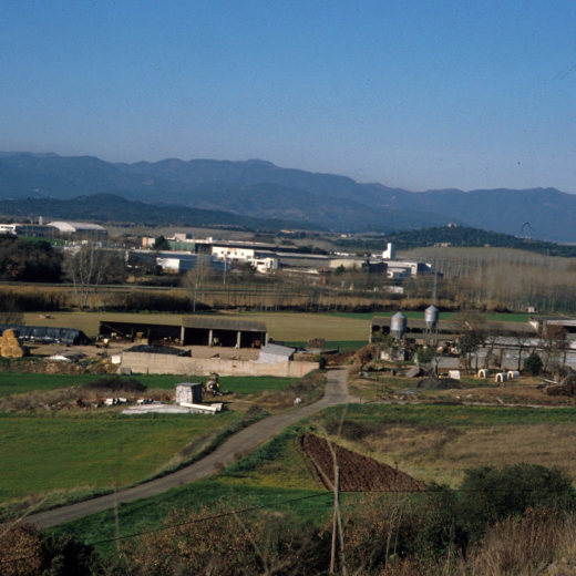 En primer terme la granja de Cal Ros i després les fàbriques del polígon industrial. Com a fons les muntanyes d'Argimon de les Guilleries i de Santa Bàrbara.