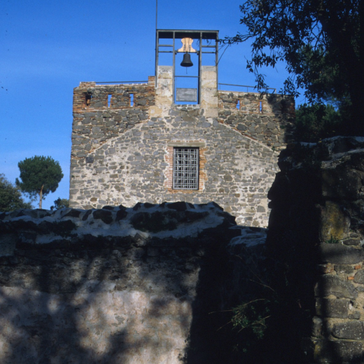 Vista de la façana principal de la Capella de Sant Jordi ja restaurada i amb la campana col·locada al nou campanar. La senyera de Catalunya presideix de dia i nit tot aquest entorn. Els veins de les cases de pagès properes es fixen amb la senyera per saber d'on bé el vent. Aquell dia tirava de tramontana.