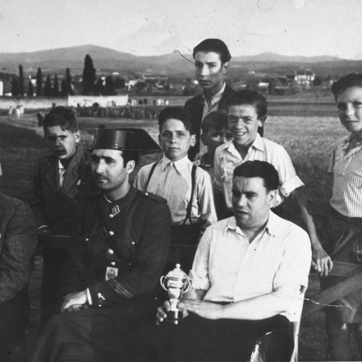 Camp de futbol de Ca l'Esparra. La presidència d'un partit de futbol per la Festa Major. El delegat de l'equip visitant, el sargent de la Guardia Civil de Vidreres i a la seva esquerra el secretari de l'Ajuntament de Maçanet, Joan Viader amb la copa o trofeu que es disputava en el partit.

A darrera hi ha els nois Jordi Costa, Joan Canals, i Josep Planas entre altres que no s'identifiquen
