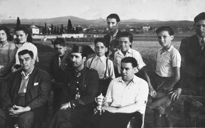 Camp de futbol de Ca l'Esparra. La presidència d'un partit de futbol per la Festa Major. El delegat de l'equip visitant, el sargent de la Guardia Civil de Vidreres i a la seva esquerra el secretari de l'Ajuntament de Maçanet, Joan Viader amb la copa o trofeu que es disputava en el partit.

A darrera hi ha els nois Jordi Costa, Joan Canals, i Josep Planas entre altres que no s'identifiquen