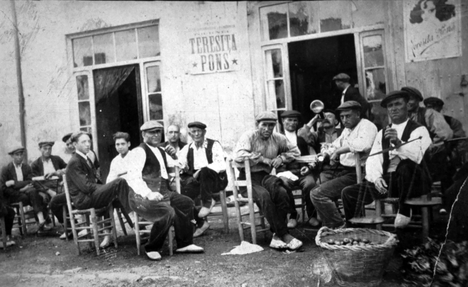 Una colla d'homes de Maçanet al davant de Can Ramón Ros, antic establiment de la plaça. Entre ells en Joan soler Pastera, Àngel Risech, Josep Ferrer, Manté amb el porró enlaire, Jaume Figueras 