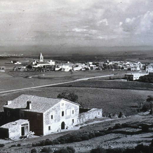 Vista de la masia Can Roura i al fons la vila de Maçanet abans de construïr-se la zona esportiva i posterior desenvolupament urbanístic.