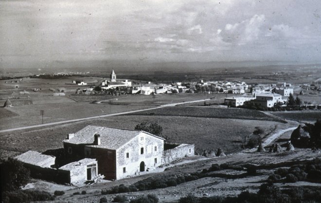 Vista de la masia Can Roura i al fons la vila de Maçanet abans de construïr-se la zona esportiva i posterior desenvolupament urbanístic.